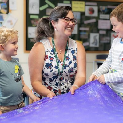 A female teacher smiles at the boy on her left. They are holding a purple sparkly cloth in front of them